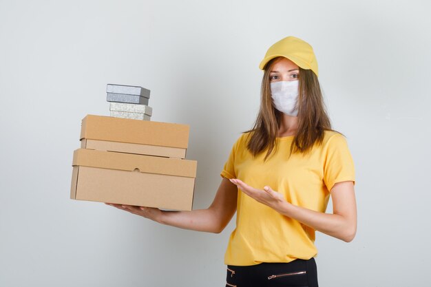 Donna delle consegne in t-shirt, pantaloni, berretto, maschera che mostra le scatole e sembra allegra