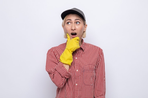 Donna delle pulizie in camicia a quadri e berretto che indossa guanti di gomma alzando lo sguardo con la mano sul mento stupita e sorpresa in piedi su sfondo bianco