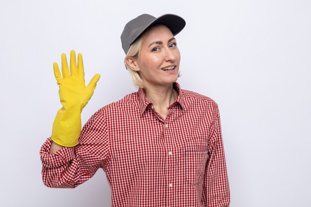 Donna delle pulizie in camicia a quadri e berretto che indossa guanti di gomma che sembra sorridente amichevole salutando con la mano