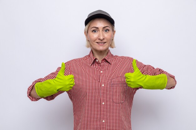 Donna delle pulizie in camicia a quadri e berretto che indossa guanti di gomma dall'aspetto felice e fiducioso sorridente che mostra i pollici in su