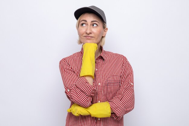 Donna delle pulizie in camicia a quadri e berretto con guanti di gomma che guarda da parte con la mano sul mento pensando positivo