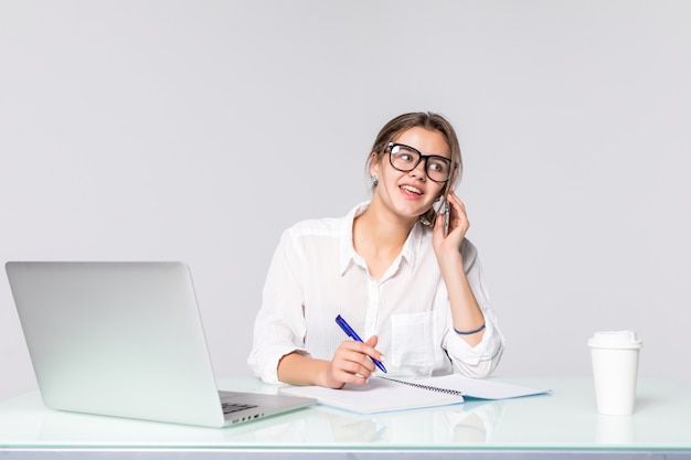 Donna di affari al suo scrittorio funzionante con il computer portatile e il telefono parlante isolati su fondo bianco