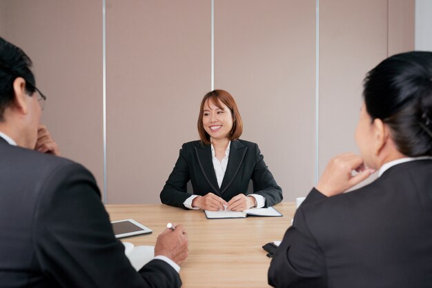 Donna di affari asiatica sicura che si siede alla riunione nell'ufficio e nel sorridere