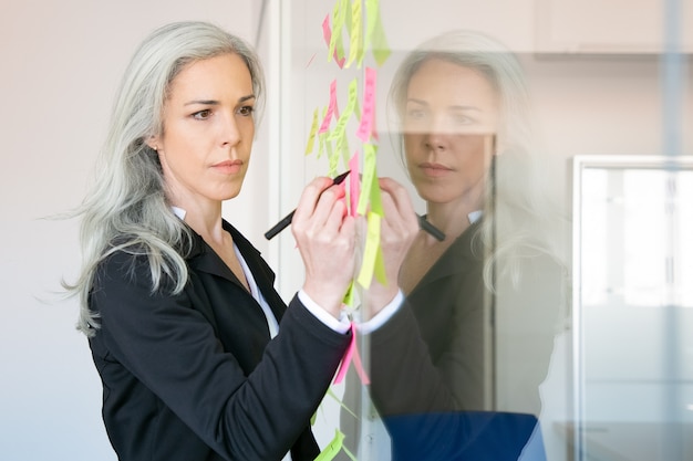 Donna di affari caucasica dai capelli grigi contenuto che scrive sull'adesivo con l'indicatore. Responsabile femminile professionale concentrato che condivide l'idea per il progetto e che prende nota.