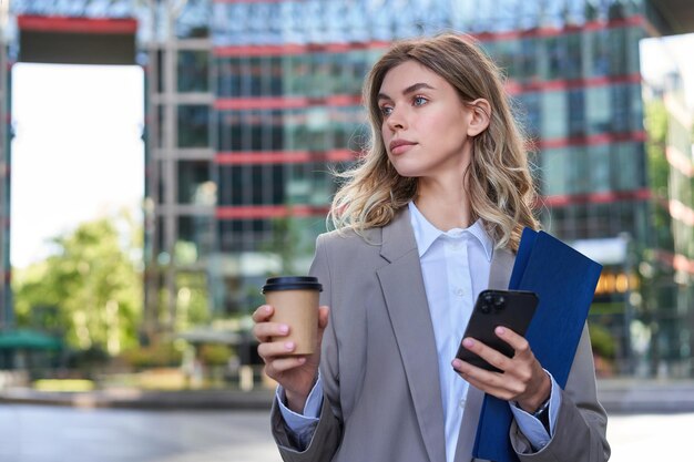 Donna di affari che beve caffè e tiene documenti con lo smartphone andando a lavorare in ufficio in piedi