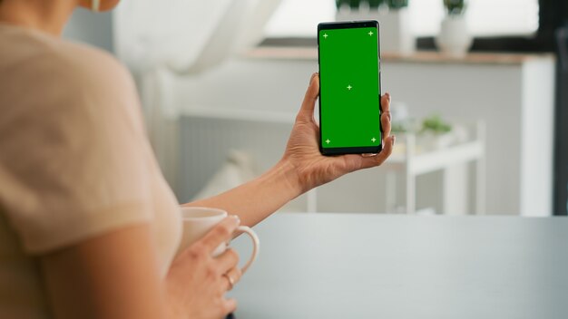 Donna di affari che naviga su internet utilizzando mock up smartphone chroma key schermo verde seduto alla scrivania dell'ufficio. Libero professionista che scorre le informazioni online per il progetto sui social media utilizzando un dispositivo isolato