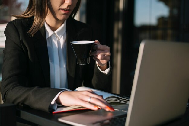 Donna di affari che per mezzo del computer portatile mentre mangiando tazza di caffè