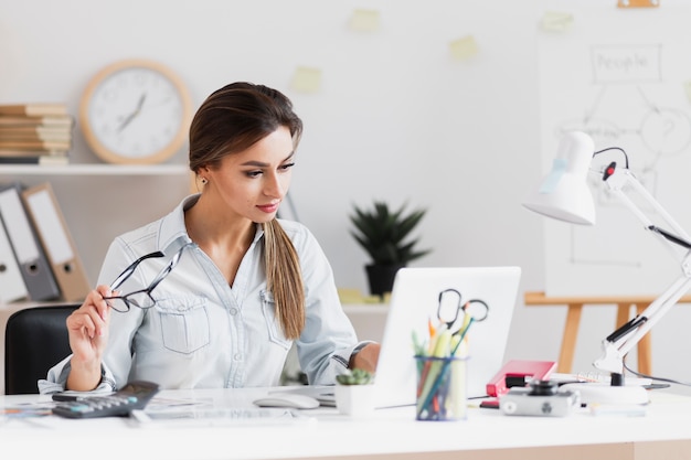 Donna di affari che tiene i suoi vetri e che lavora al computer portatile