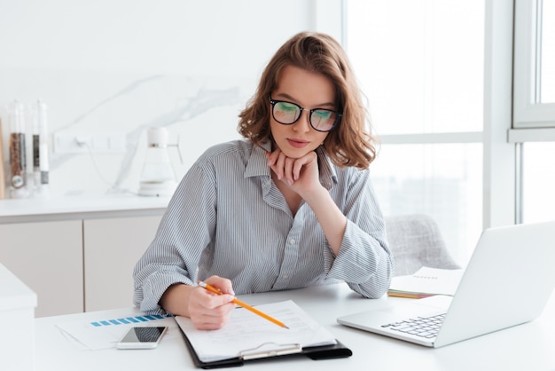 Donna di affari concentrata giovani in vetri e camicia a strisce che lavorano con le carte a casa