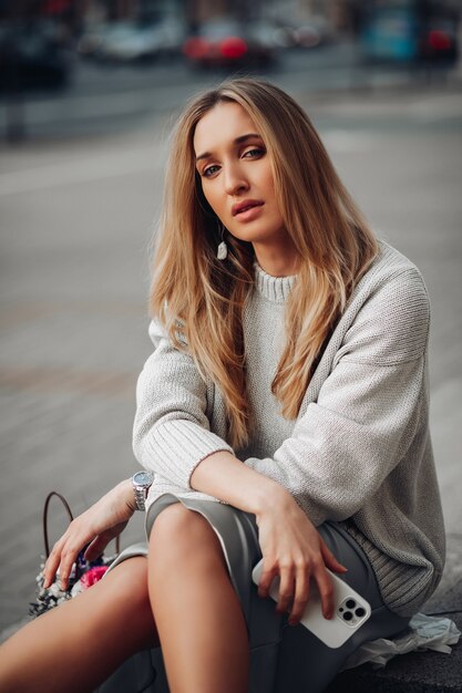 Donna di affari della ragazza alla moda abbastanza giovane con la seduta del telefono, posante sulla via. Moda femminile, stile casual e femminilità. Foto di alta qualità