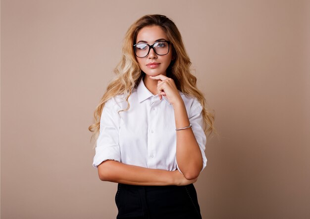 Donna di affari in occhiali alla moda che controllano parete beige. Capelli biondi ondulati Camicetta bianca e pantaloni neri.