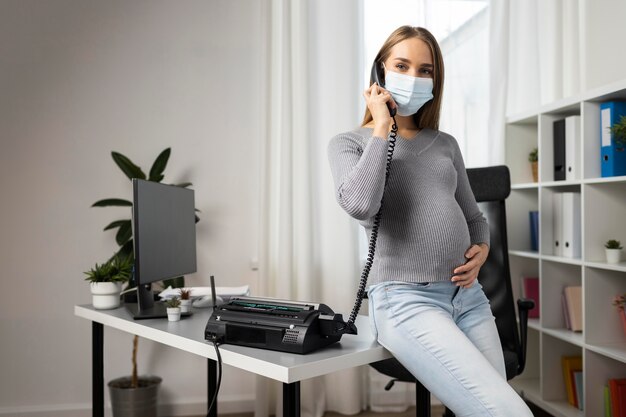 Donna di affari incinta che prende le chiamate in ufficio mentre indossa la mascherina medica