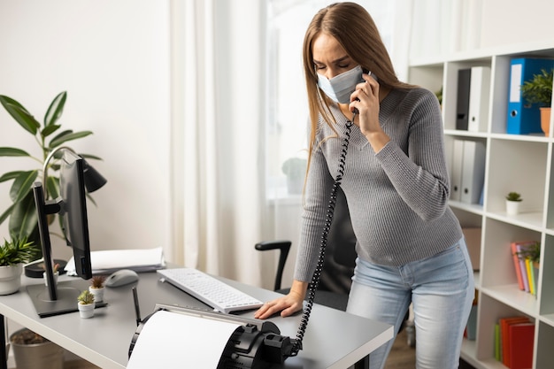 Donna di affari incinta con mascherina medica che risponde alle chiamate in ufficio