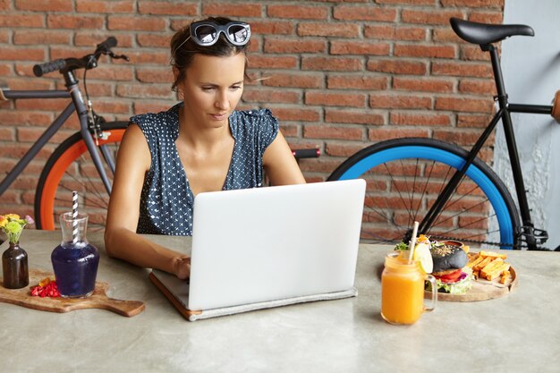 Donna di affari seria con le tonalità sulla sua testa che controlla email sul suo computer portatile moderno durante il pranzo nel fine settimana. Lavoratrice autonoma che utilizza un notebook per lavoro a distanza