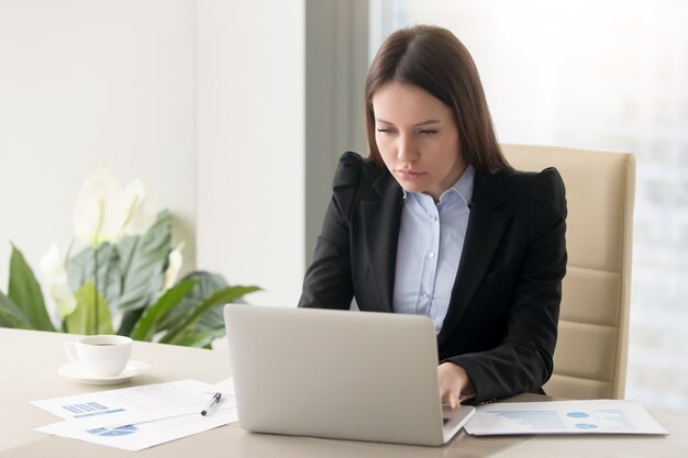 Donna di affari seria concentrata che stendere rapporto, funzionante con il computer portatile in ufficio