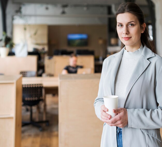 Donna di affari sorridente che tiene la tazza di caffè eliminabile nel posto di lavoro