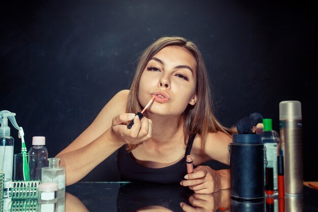 Donna di bellezza che applica trucco. Bella ragazza guardarsi allo specchio e applicare cosmetici con un pennello. Mattina, trucco e concetto di emozioni umane. Modello caucasico in studio