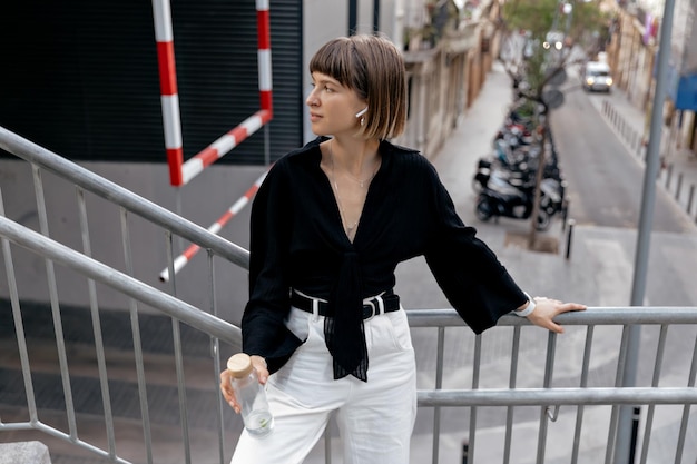 Donna di buonumore in piedi fuori Ragazza dai capelli corti in camicia nera e pantaloni bianchi in cuffie wireless con in mano una bottiglia d'acqua e sorrisi all'aperto