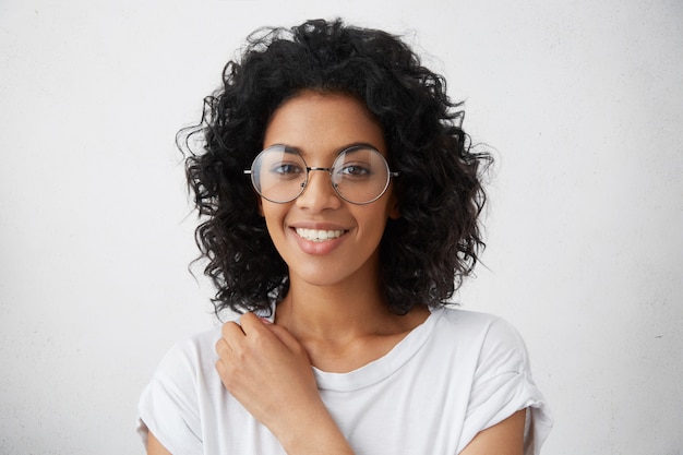 Donna di college nero di razza mista con acconciatura riccia che sorride ampiamente vestita in camicetta bianca, guardando fresca e giovane, indossando occhiali alla moda. Emozioni e sentimenti umani positivi