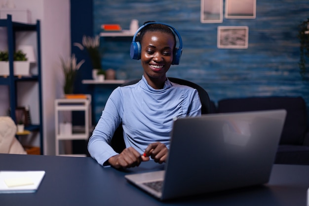 Donna di colore di buon umore che indossa l'auricolare che ascolta musica lavorando alla scadenza dall'ufficio a casa. seduto alla scrivania. Libero professionista africano che crea un nuovo progetto lavorando fino a tardi.