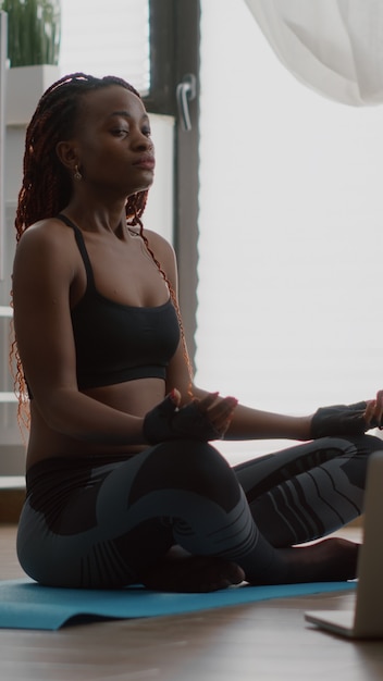 Donna di colore esile che si siede nella posa del loto sulla mappa di yoga con gli occhi chiusi