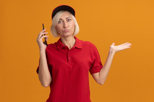 Donna di consegna bionda di mezza età confusa in uniforme rossa e berretto guardando davanti tenendo il telefono cellulare vicino alla testa che mostra la mano vuota isolata sulla parete arancione