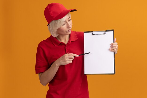 Donna di consegna bionda di mezza età impressionata in uniforme rossa e cappuccio che indica la lavagna per appunti con la matita che la esamina isolata sulla parete arancione con lo spazio della copia