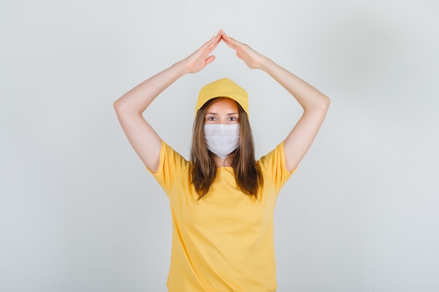 Donna di consegna che fa il segno del tetto della casa sopra la testa in t-shirt, berretto, maschera e sembra felice