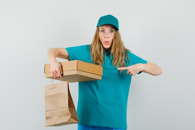 Donna di consegna che indica alla scatola di cartone e che tiene il sacchetto di carta in maglietta, berretto e che sembra curiosa.