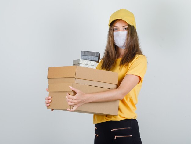 Donna di consegna che tiene cartone e scatole presenti in t-shirt, pantaloni e berretto, maschera e aspetto allegro