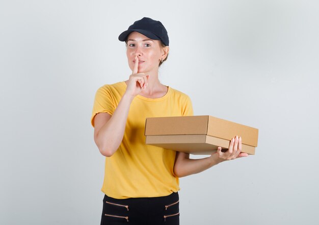 Donna di consegna che tiene la scatola di cartone con gesto di silenzio in t-shirt, pantaloni e berretto