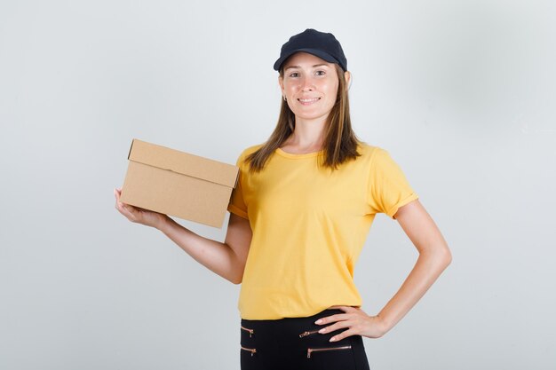 Donna di consegna che tiene la scatola di cartone in t-shirt, pantaloni e berretto e sembra allegra