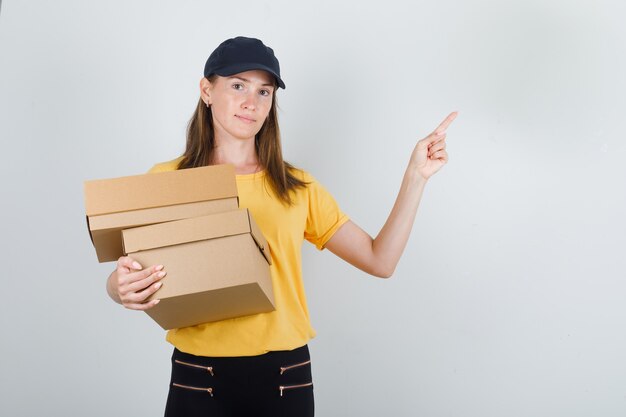 Donna di consegna che tiene le scatole di cartone con il dito in su in maglietta, pantaloni e cappello