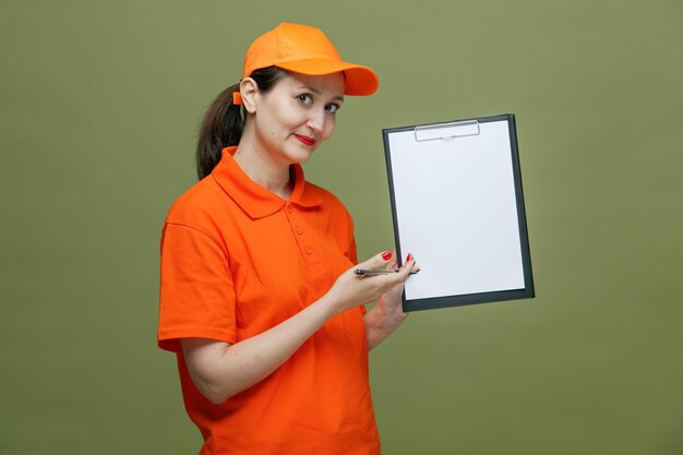 Donna di consegna di mezza età impressionata che indossa l'uniforme e la penna che tiene il cappuccio guardando la fotocamera che mostra gli appunti isolati su sfondo verde oliva