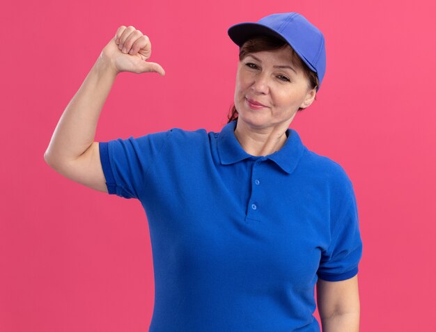 Donna di consegna di mezza età in uniforme blu e berretto che guarda davanti con un'espressione fiduciosa che punta a se stessa in piedi sopra il muro rosa