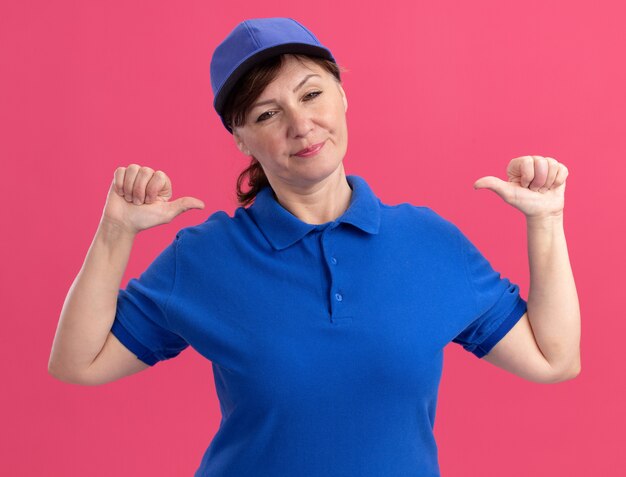 Donna di consegna di mezza età in uniforme blu e berretto guardando la parte anteriore che punta a se stessa in piedi sul muro rosa