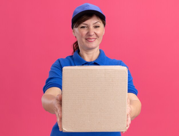 Donna di consegna di mezza età in uniforme blu e cappuccio che mostra la scatola di cartone guardando davanti sorridente fiducioso in piedi sopra il muro rosa