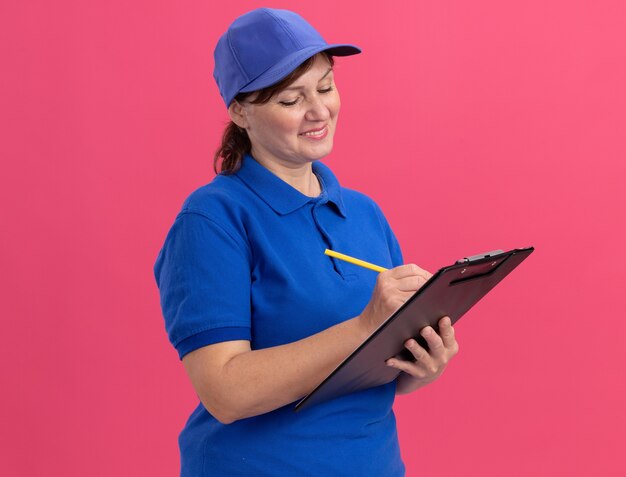 Donna di consegna di mezza età in uniforme blu e cappuccio che tiene appunti e scrittura a matita con il sorriso sul viso in piedi sopra il muro rosa
