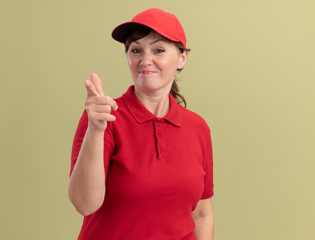 Donna di consegna di mezza età in uniforme rossa e berretto che sembra fiducioso che punta con il dito indice davanti in piedi sopra la parete verde