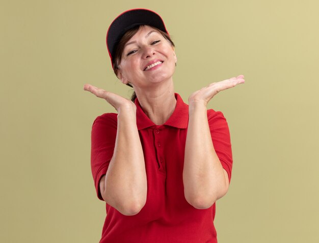 Donna di consegna di mezza età in uniforme rossa e berretto guardando davanti sorridente felice e positivo con le mani alzate in piedi sopra la parete verde