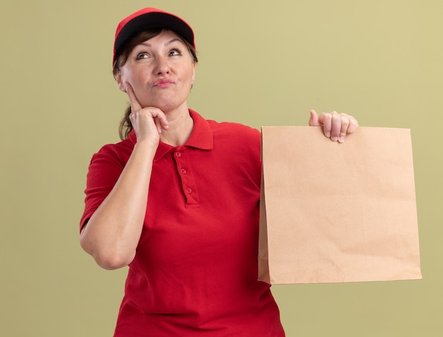 Donna di consegna di mezza età in uniforme rossa e cappuccio che dà pacchetto di carta che osserva in su con espressione pensierosa sul pensiero del viso in piedi sopra la parete verde