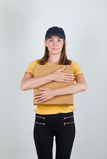 Donna di consegna in t-shirt, pantaloni, berretto che abbraccia la scatola di cartone e sembra contenta