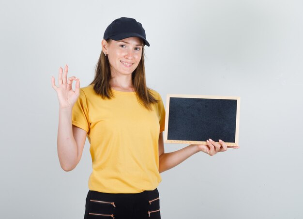 Donna di consegna in t-shirt, pantaloni, berretto che tiene lavagna con segno ok e sembra contenta