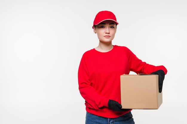 Donna di consegna in uniforme rossa isolata sulla parete bianca. Corriere in guanti medicali, berretto, maglietta rossa che lavora come rivenditore che tiene una scatola di cartone da consegnare. Pacchetto di ricezione.