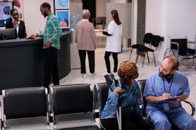 Donna di consulenza specialistica sanitaria con stampelle nella hall della reception dell'ospedale, che parla di diagnosi e medicina nell'area della sala d'attesa. Diverse persone che discutono all'appuntamento di controllo.