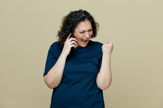 Donna di mezza età arrabbiata che indossa una maglietta che guarda in basso tenendo il pugno in aria gridando mentre parla al telefono isolato su sfondo verde oliva
