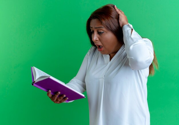 Donna di mezza età caucasica casuale sorpresa che tiene e che esamina il libro e che mette la mano sulla testa isolata sulla parete verde