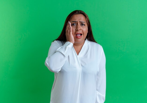 Donna di mezza età caucasica casuale spaventata che mette la mano sulla guancia isolata sulla parete verde