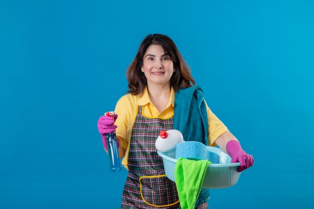 Donna di mezza età che indossa un grembiule e guanti di gomma che tengono spray per la pulizia e bacino con strumenti di pulizia