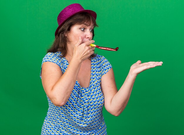 Donna di mezza età con cappello da festa che fischia felice e allegra che celebra la festa di compleanno in piedi sul muro verde